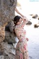 A woman in a floral dress leaning against a rock by the water.