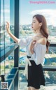 A woman in a white blouse and black skirt standing by a window.